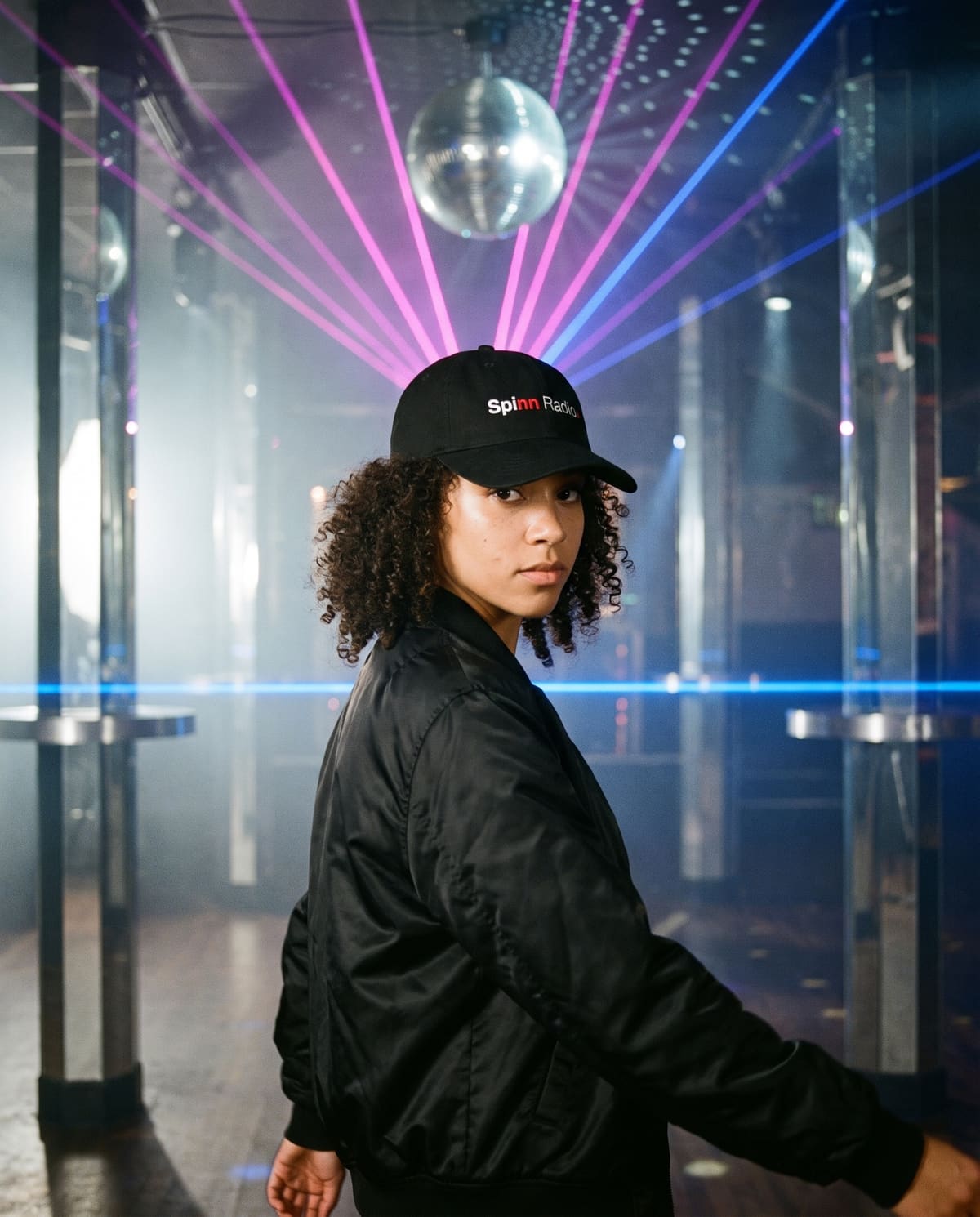 A dancer under a mirror ball in a chrome-and-haze club, wearing the black Spinn Radio cap