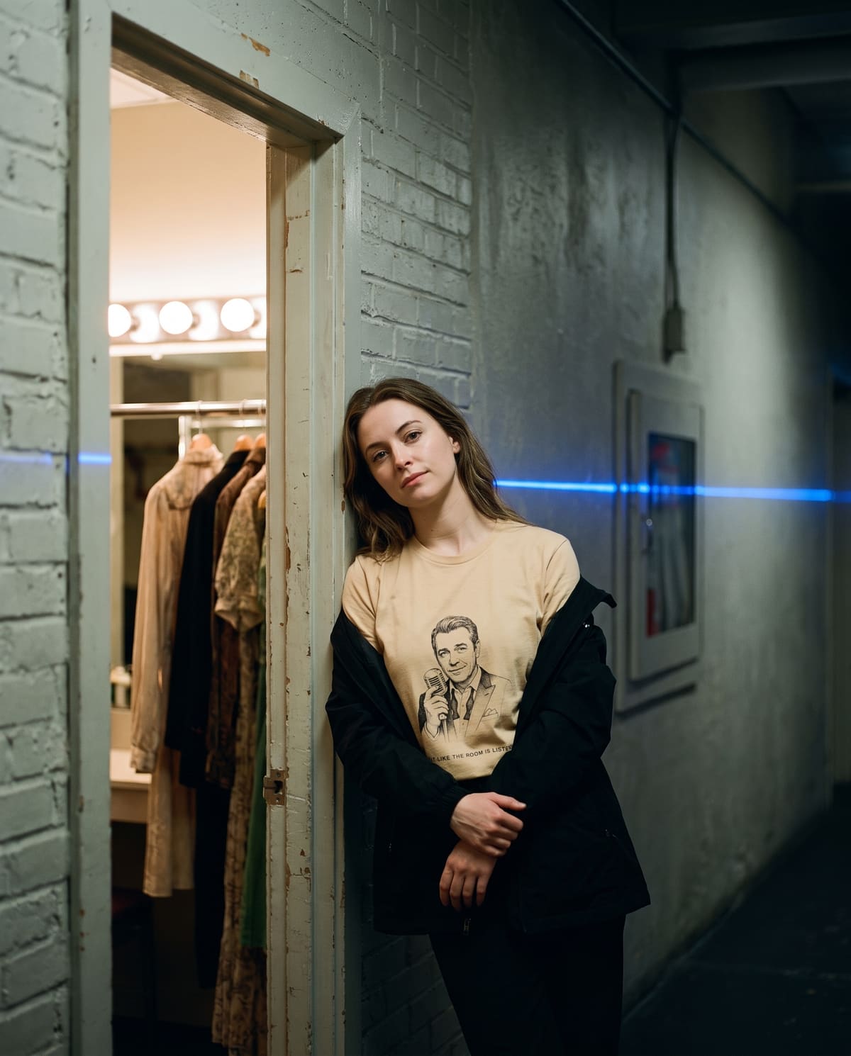 A woman leaning in a dressing-room doorway backstage, wearing the Crooner tee under an open jacket, bulb mirror lights behind her