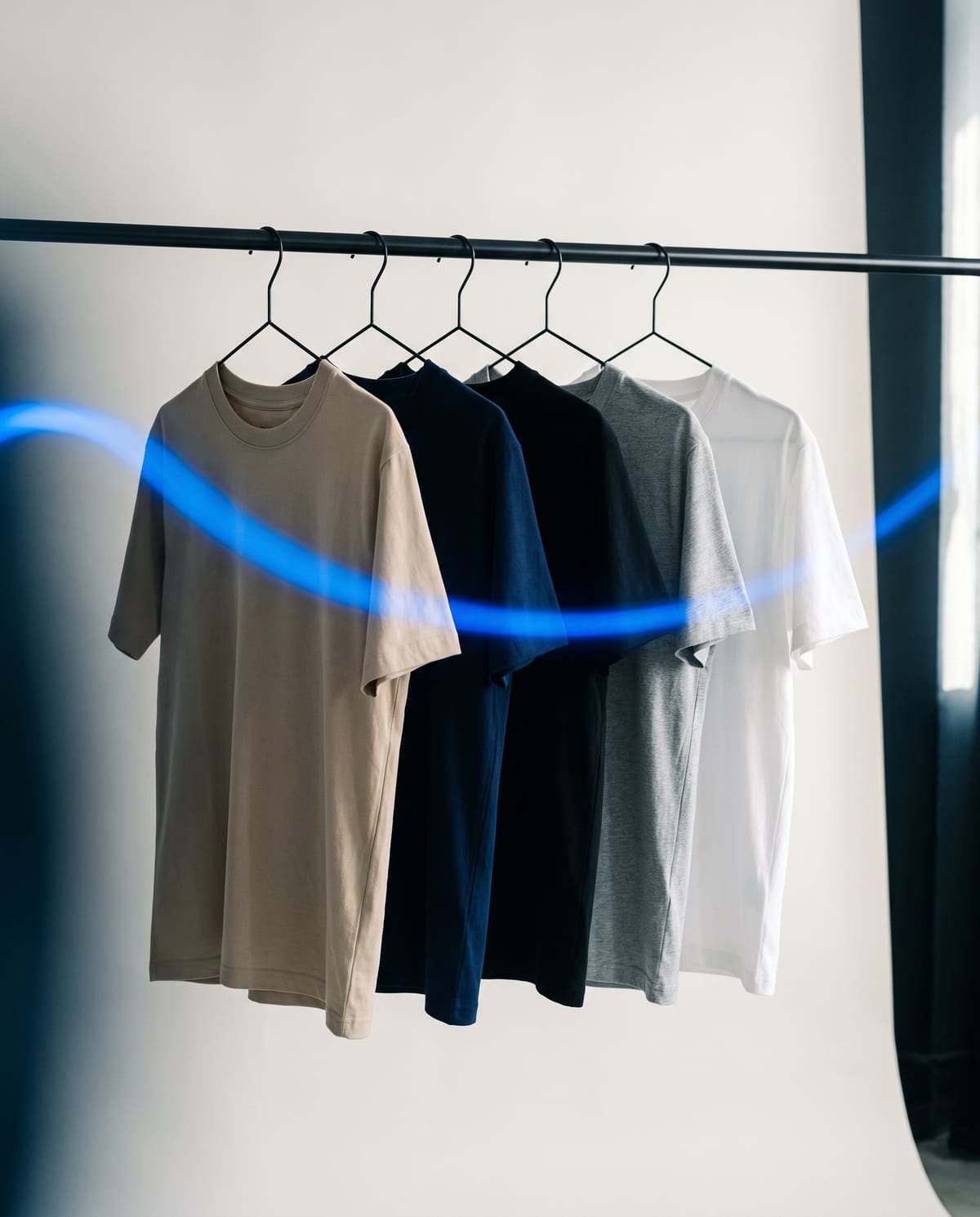 Row of blank tees in sand, navy, black and white on a rail in a bone-white studio