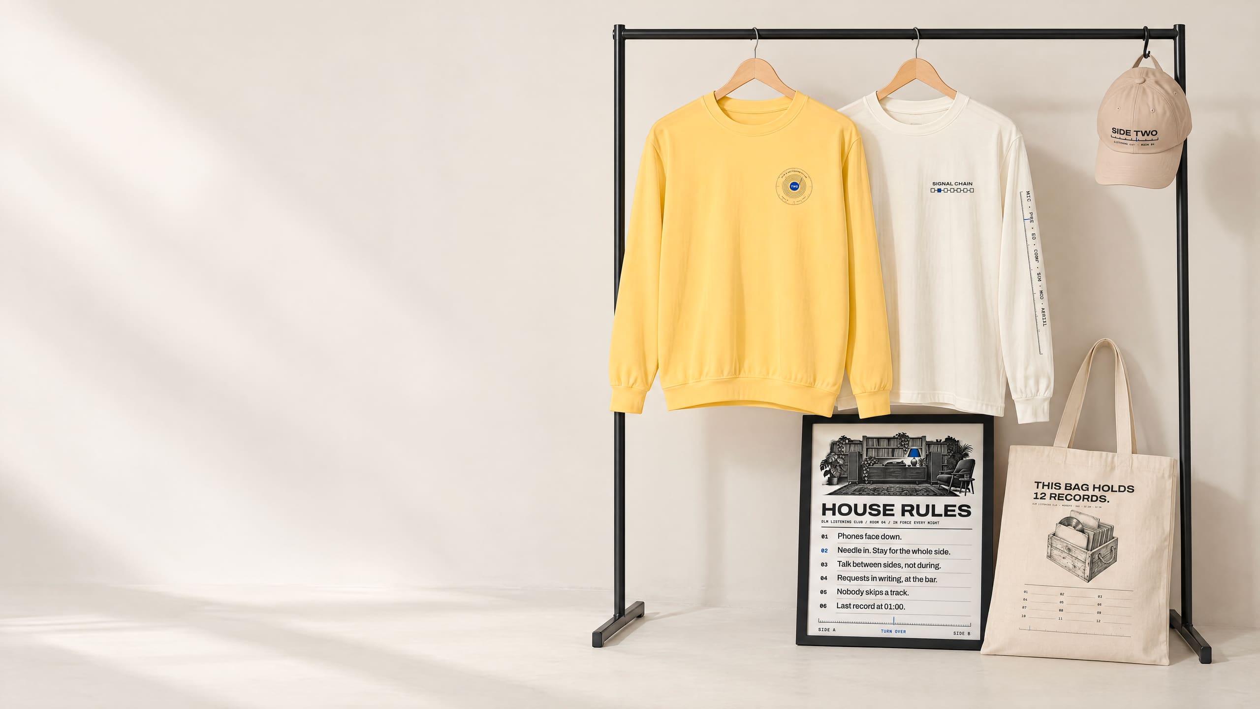 The new range on a black clothing rail in a bone-white studio: a butter-yellow crewneck, an ivory long sleeve, a stone cap, a framed House Rules print and a canvas tote